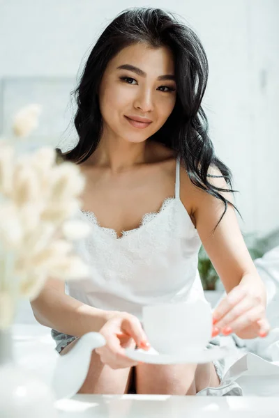 Selective focus of happy brunette thai woman holding saucer and cup of tea — Stock Photo