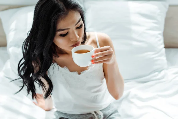 Selektiver Fokus der brünetten Thailänderin, die im Schlafzimmer eine Tasse mit Tee hält — Stockfoto