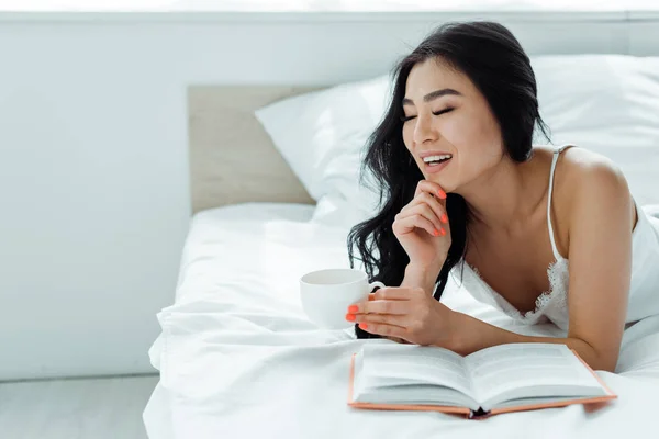Selective focus of cheerful brunette asian woman lying near book and holding cup of tea — Stock Photo
