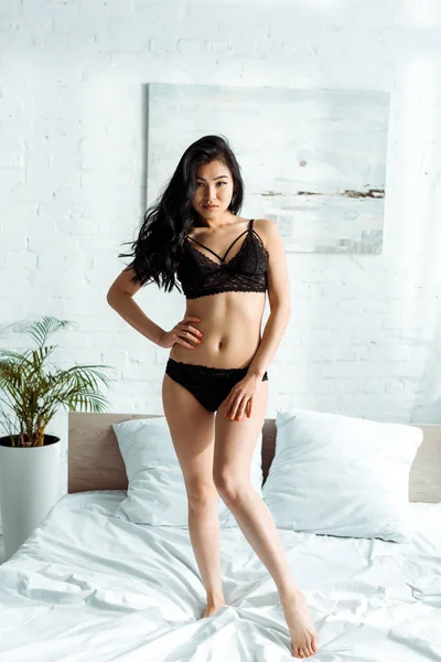 Чувственная азиатская молодая женщина смотрит в камеру и стоит на кровати — стоковое фото