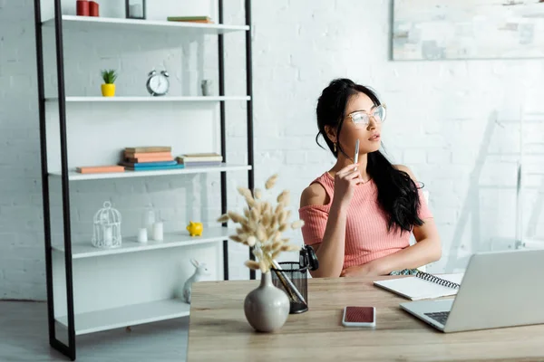 Atractivo asiático mujer en gafas sentado en oficina cerca de portátil - foto de stock