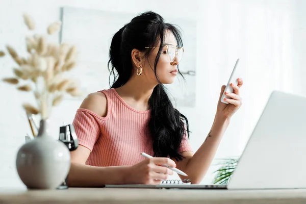 Selektiver Fokus einer asiatischen Frau mit Brille, die im Büro auf ihr Smartphone blickt — Stockfoto