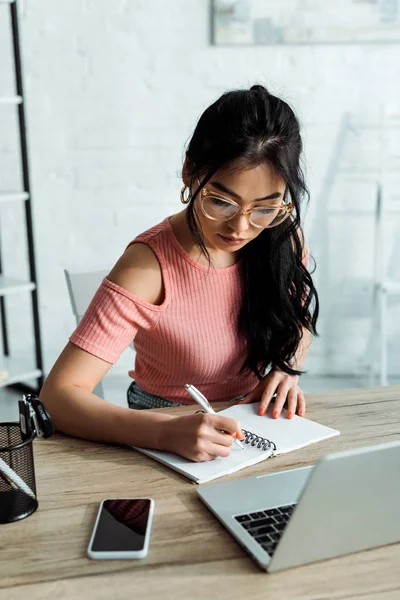 Attractive thai woman in glasses writing in notebook near gadgets — Stock Photo