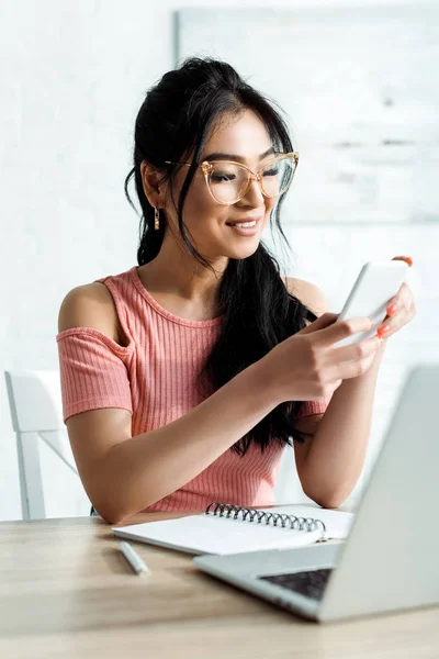 Enfoque selectivo de la mujer asiática feliz en gafas usando teléfono inteligente cerca de la computadora portátil en la oficina - foto de stock