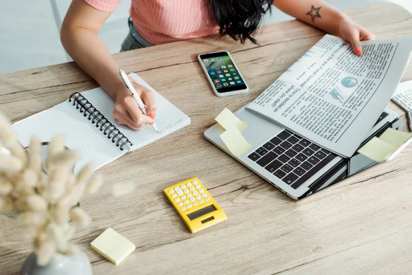 KYIV, UCRAINA - 23 MAGGIO 2019: vista ritagliata della donna che scrive sul taccuino vicino al giornale e all'iphone — Foto stock