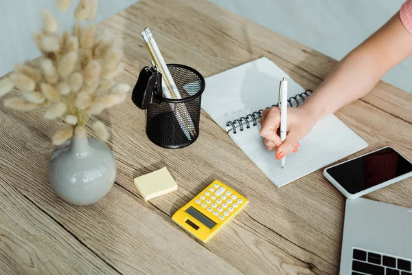 Vista ritagliata di donna che scrive in notebook vicino a giornale e gadget — Foto stock