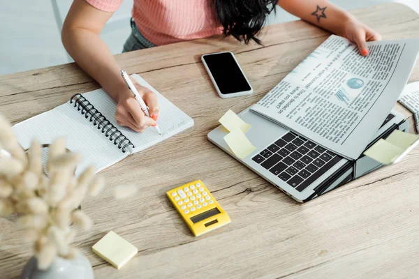 Ausgeschnittene Ansicht einer jungen Frau, die in Notizbuch neben Zeitung und Gadgets schreibt — Stockfoto