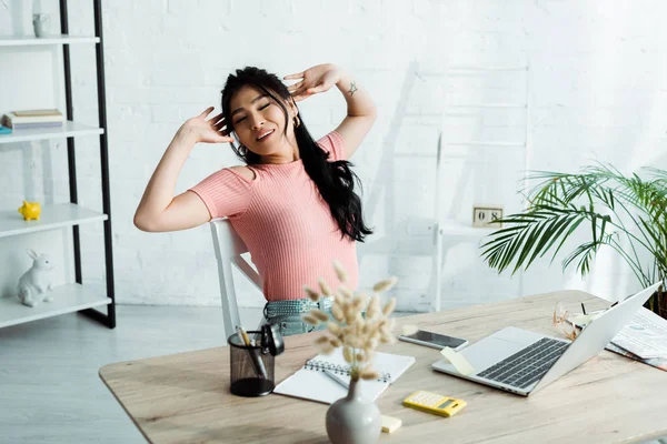 Selective focus of attractive asian woman with closed eyes stretching while sitting near table — Stock Photo