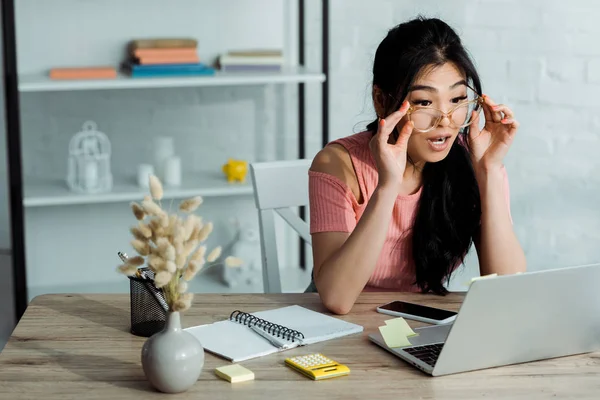 Enfoque selectivo de mujer asiática sorprendida tocando gafas y mirando a la computadora portátil - foto de stock
