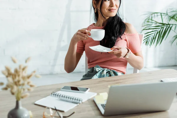 Selective focus of cheerful woman holding cup with drink and saucer near gadgets — Stock Photo