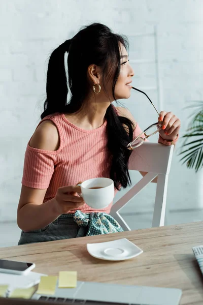 Selective focus of happy young asian woman holding gasses near lips and cup of tea — Stock Photo