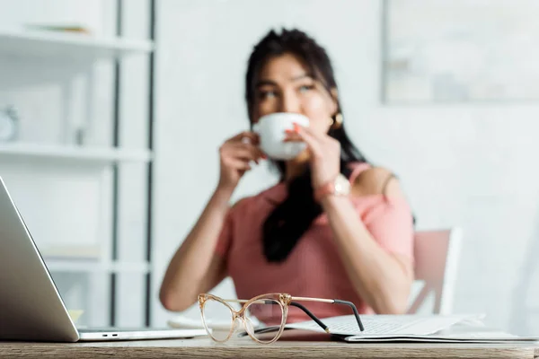Fuoco selettivo di occhiali sul tavolo vicino donna asiatica bere il tè — Foto stock