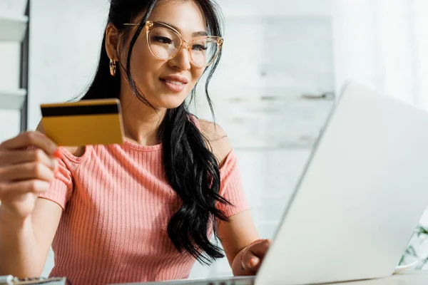 Selective focus of cheerful thai woman in glasses using laptop while holding credit card — Stock Photo