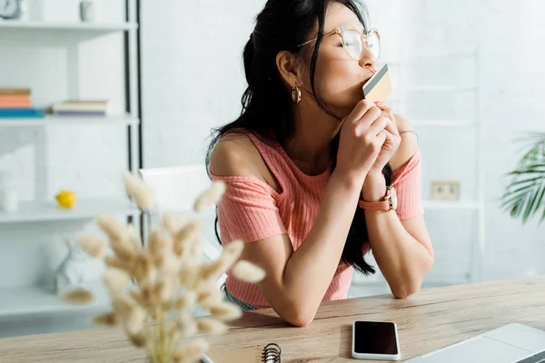 Enfoque selectivo de atractiva mujer asiática con ojos cerrados besos tarjeta de crédito cerca de teléfono inteligente con pantalla en blanco - foto de stock