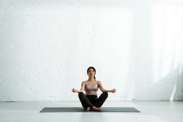Mujer tailandesa tranquila haciendo ejercicio mientras está sentada con las piernas cruzadas en la esterilla de yoga - foto de stock