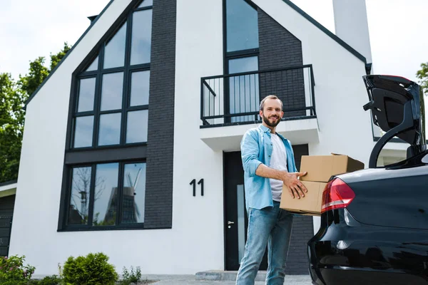 Vista de ángulo bajo del hombre alegre que sostiene cajas cerca del coche y de la casa moderna - foto de stock