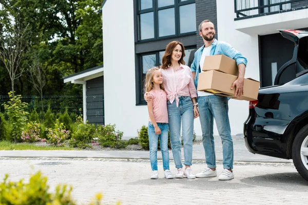 Cheerful bearded man holding boxes near happy wife and daughter — Stock Photo