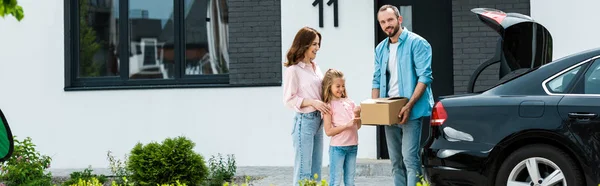 Panoramic shot of cheerful family moving into modern house while standing near car — Stock Photo