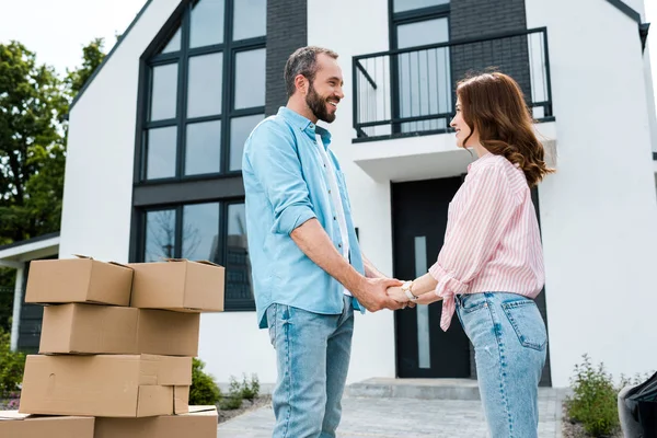Happy woman holding hands with bearded man near boxes and new house — Stock Photo