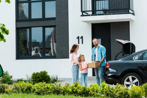 Hombre feliz sosteniendo caja cerca de esposa e hija mientras está de pie cerca del coche - foto de stock