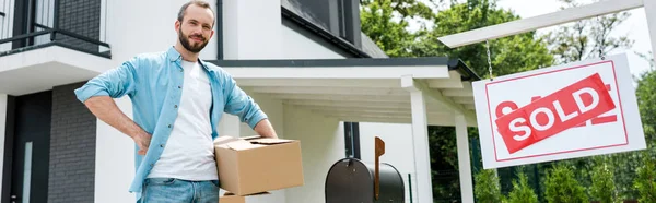 Panoramic shot of handsome man standing with hand on hip and holding box near new house and board with sold letters — Stock Photo