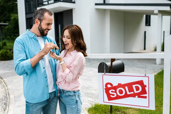 Cheerful man holding key and standing with happy woman near board with sold letters — Stock Photo