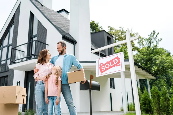 Bearded man holding box and standing with wife and daughter near house and board with sold letters — Stock Photo