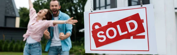 Panoramic shot of board with sold lettering near happy man gesturing with woman — Stock Photo