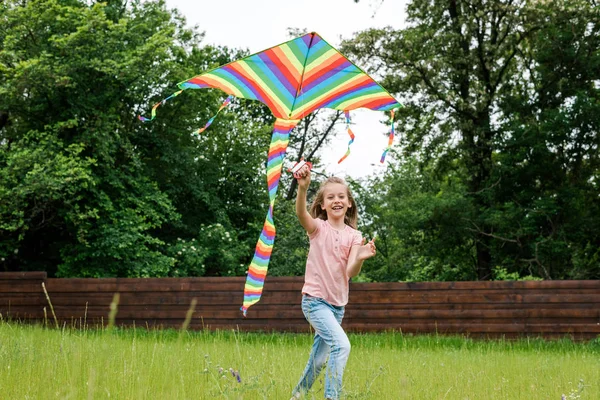 Glückliches Kind läuft mit buntem Drachen auf grünem Gras draußen — Stockfoto