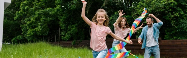 Tiro panorámico de niño feliz corriendo con cometa colorida cerca de padres alegres - foto de stock