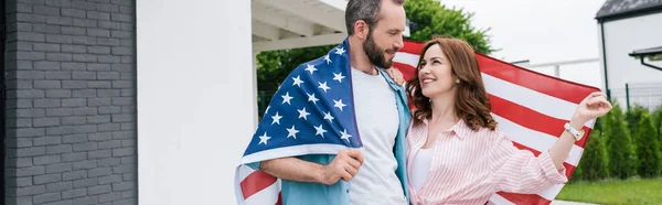 Panoramic shot of happy bearded man standing with attractive woman and holding american flag — Stock Photo