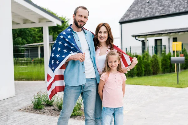 Famille heureuse debout avec drapeau américain près de la maison — Photo de stock
