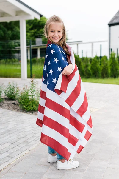 Glückliches Kind lächelt, während es mit amerikanischer Flagge in der Nähe des Hauses steht — Stockfoto
