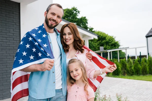 Niño feliz cerca de padres alegres con bandera americana de pie cerca de la casa - foto de stock