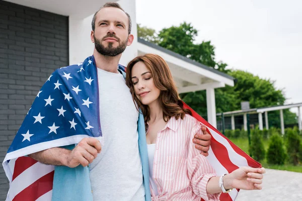 Atractiva mujer con los ojos cerrados sosteniendo la bandera americana y de pie con el hombre guapo - foto de stock