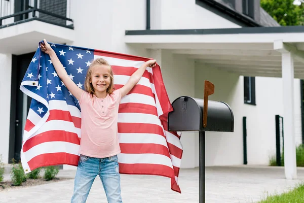 Happy kid standing and holding american flag near mail box and house — Stock Photo
