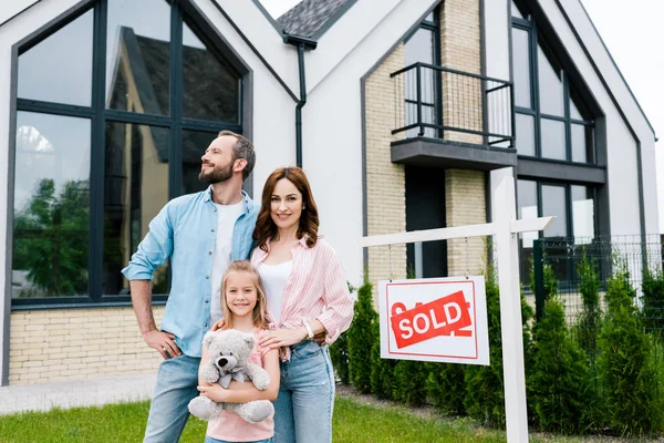 Alegre niño sosteniendo osito de peluche y de pie con los padres cerca de casa y vende letras a bordo - foto de stock