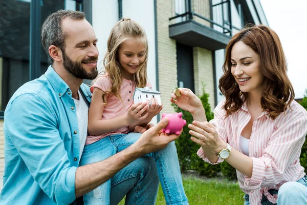 Happy woman putting golden coin in piggy bank near husband and daughter — Stock Photo