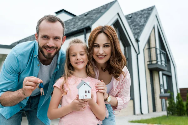 Niño alegre sosteniendo modelo de la casa de cartón cerca del padre con llaves y madre feliz - foto de stock