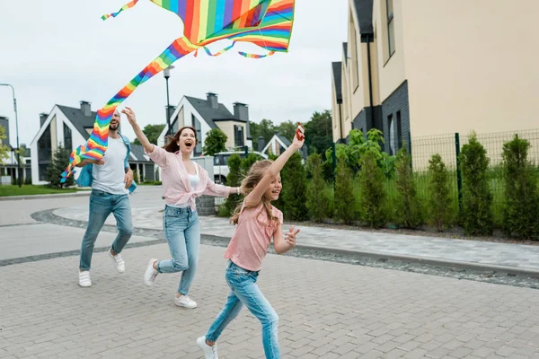 Niño feliz corriendo con colorido cometa cerca de padres alegres en la calle - foto de stock