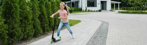 Plan panoramique d'enfant heureux souriant tout en vélo scooter près des arbres dans la rue — Photo de stock