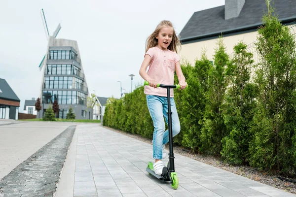 Low angle view of cute kid riding scooter near trees on street — Stock Photo