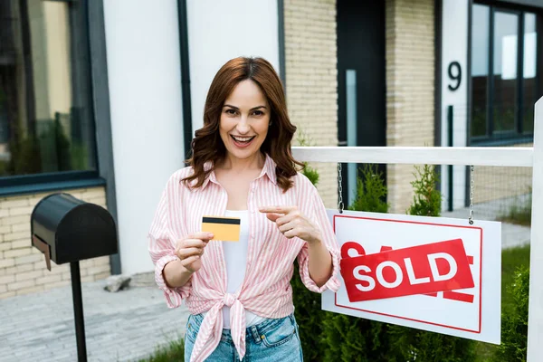 Femme gaie pointant avec le doigt à la carte de crédit près de bord avec des lettres vendues — Photo de stock