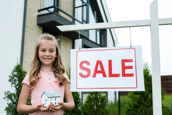 Niño alegre sosteniendo el modelo de la casa de cartón cerca del tablero con las letras de la venta - foto de stock
