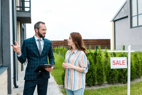 Handsome bearded broker holding clipboard and gesturing near attractive woman and board with sale letters — Stock Photo