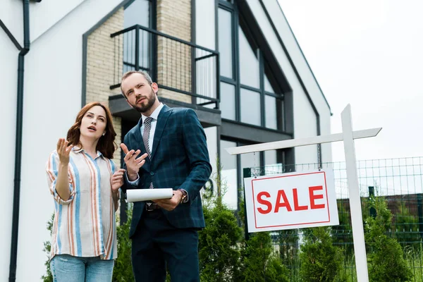 Handsome broker holding clipboard and gesturing near attractive woman and board with sale letters — Stock Photo