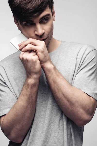 Hombre pensativo mirando hacia otro lado mientras sostiene el teléfono inteligente cerca de la cara aislado en gris, emoción humana y concepto de expresión - foto de stock