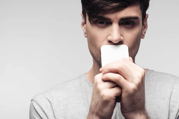 Серьезный, вдумчивый человек, держащий смартфон рядом с лицом, изолированным от серого — стоковое фото