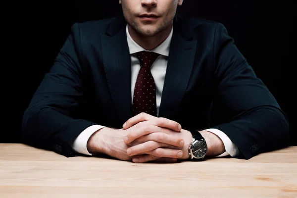 Visión parcial del hombre de negocios sentado en la mesa con las manos apretadas aisladas en negro, emoción humana y concepto de expresión - foto de stock