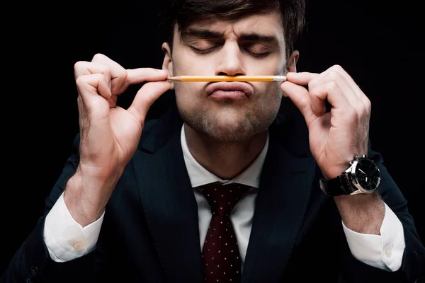 Joven hombre de negocios con los ojos cerrados haciendo muecas mientras sostiene el lápiz cerca de la nariz aislado en negro - foto de stock